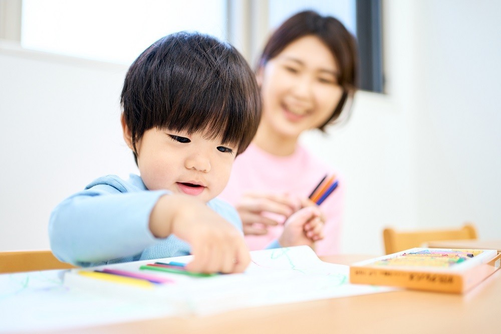お絵描きをしている男の子を後ろから見守る保育士