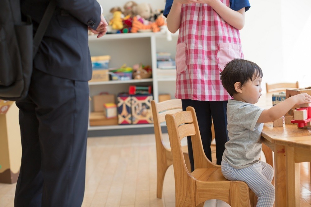 椅子に座って遊ぶ子どもの後ろで話をする保育士と保護者の足元